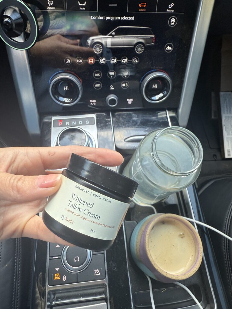 A hand holds a jar labeled Whipped Tallow Cream above a car&rsquo;s center console. Nearby are an empty glass jar, a ceramic mug, and white charging cables resting on the console.