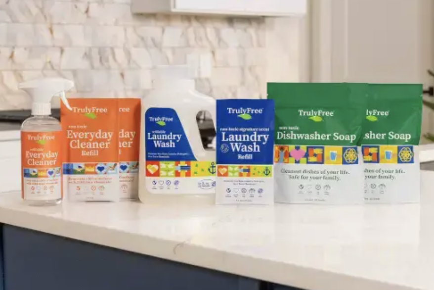 A row of TrulyFree cleaning products, including Everyday Cleaner, Laundry Wash, and Dishwasher Soap, displayed on a white kitchen counter with a tiled backsplash in the background.
