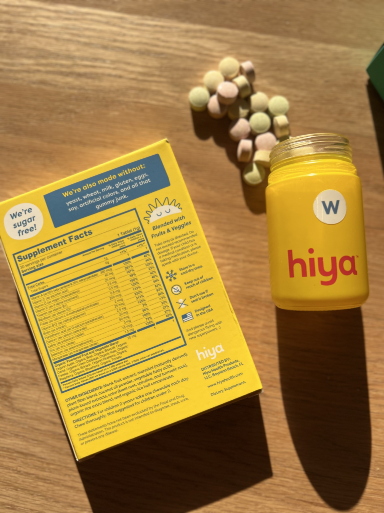 A yellow bottle of Hiya vitamins stands next to its open box on a wooden surface. Scattered around the bottle are pastel-colored vitamin tablets.