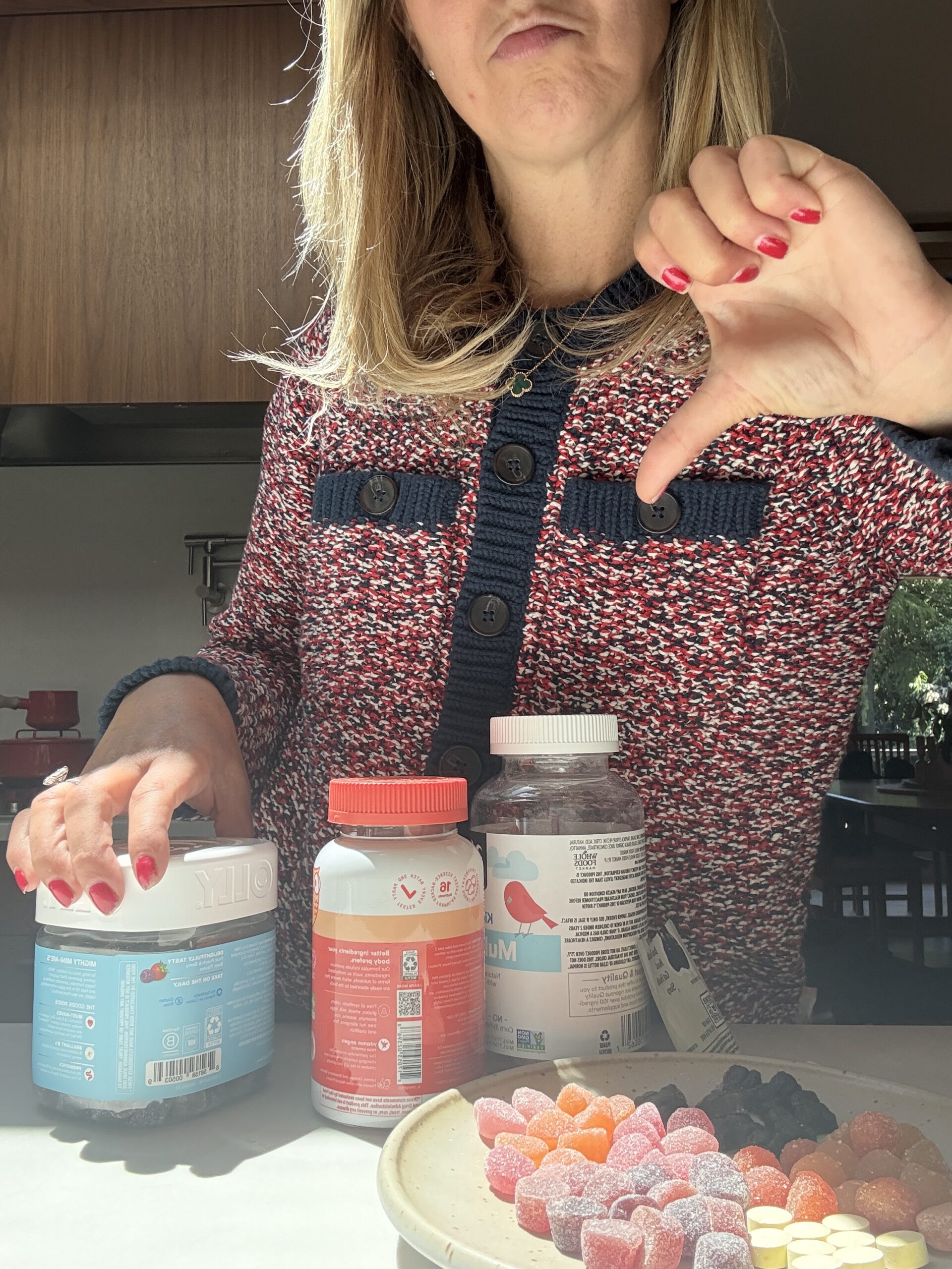 A woman with long blond hair and red nail polish gives a thumbs-down gesture while standing in front of containers of vitamins and a plate with assorted gummy supplements on a kitchen counter.