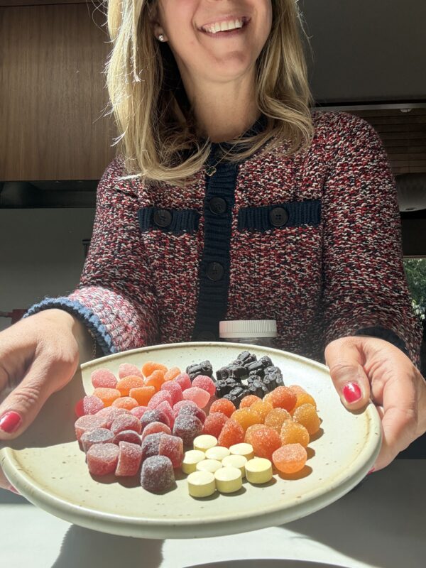 A woman in a patterned sweater smiles while holding a plate with assorted colorful gummies and round candies, offering them forward in bright sunlight.