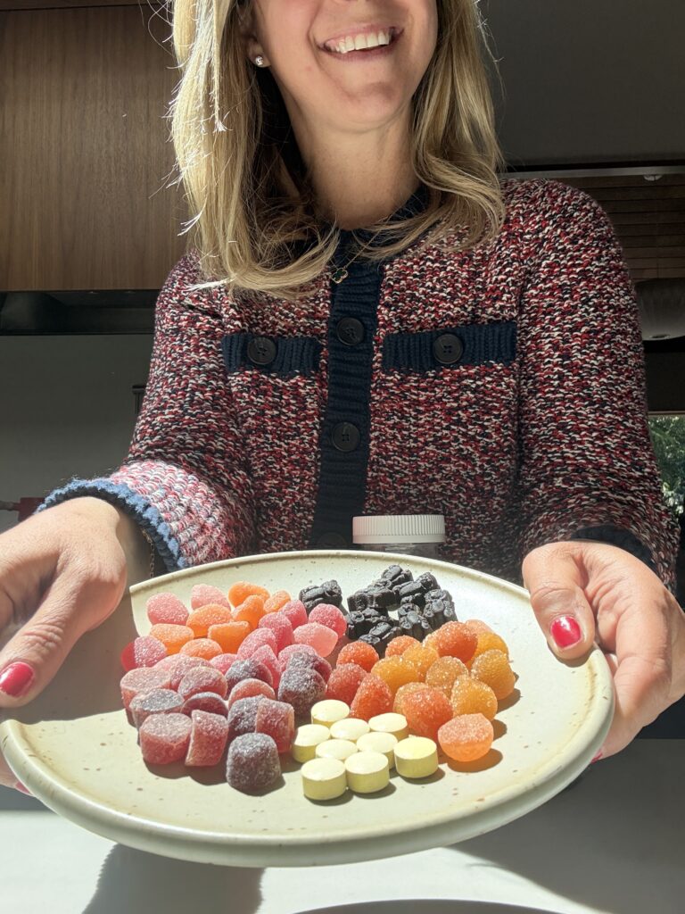 A woman in a patterned sweater smiles while holding a plate with assorted colorful gummies and round candies, offering them forward in bright sunlight.