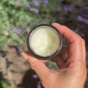 A hand holds an open jar of creamy white substance, possibly lotion or balm, against a blurred outdoor background with green foliage and purple flowers.