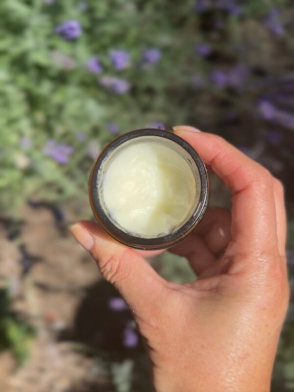 A hand holds an open jar of creamy white substance, possibly lotion or balm, against a blurred outdoor background with green foliage and purple flowers.