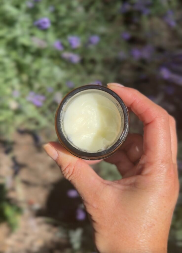 A hand holds an open jar of creamy white substance, possibly lotion or balm, against a blurred outdoor background with green foliage and purple flowers.