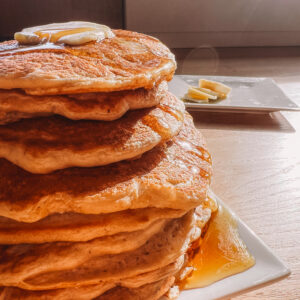 A stack of pancakes with butter and syrup sits on a white plate, with a few banana slices on another plate in the background in sunlight.