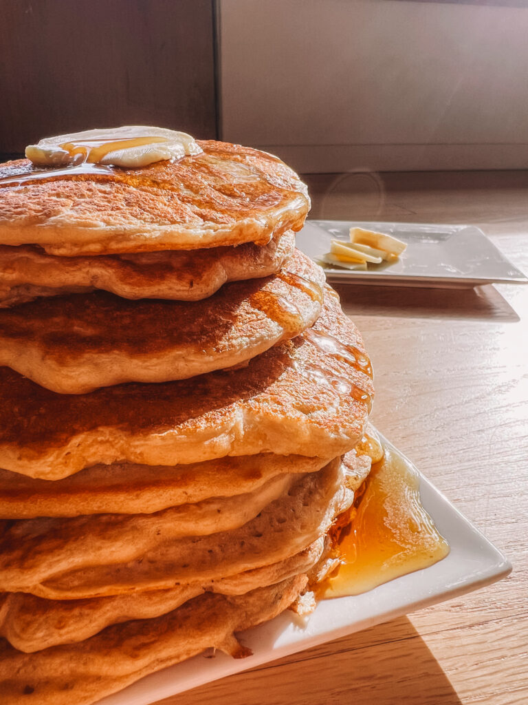 A stack of pancakes with butter and syrup sits on a white plate, with a few banana slices on another plate in the background in sunlight.