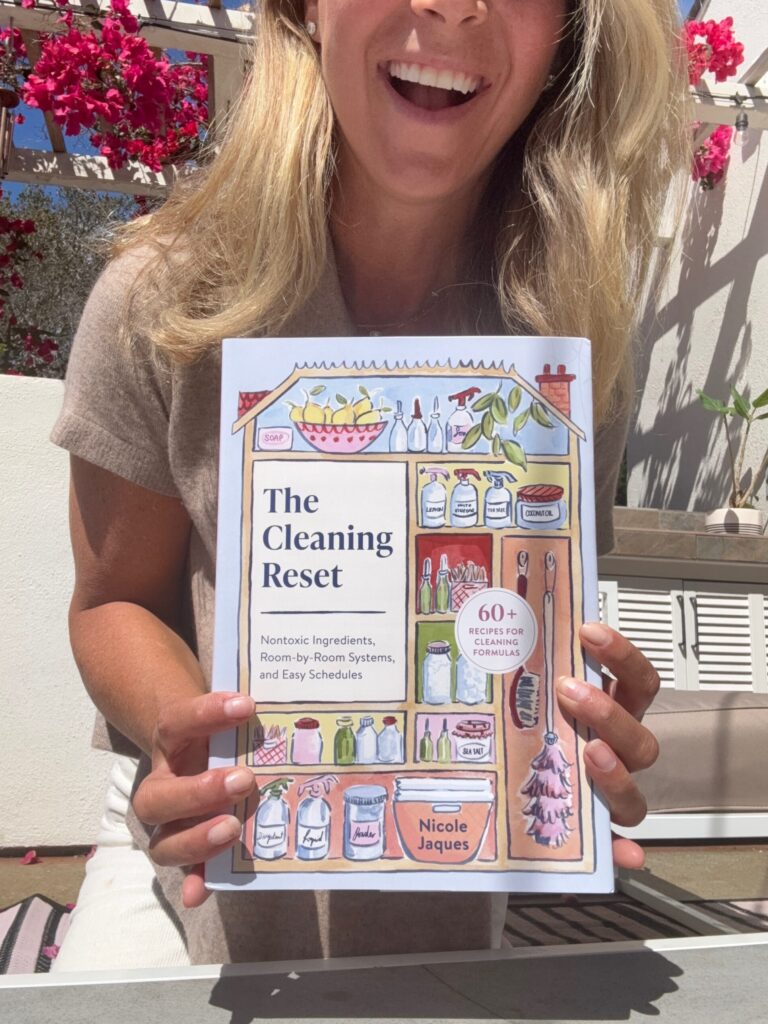A woman smiling outdoors holds a book titled The Cleaning Recer with illustrated cleaning supplies on the cover; pink flowers and sunlight appear in the background.